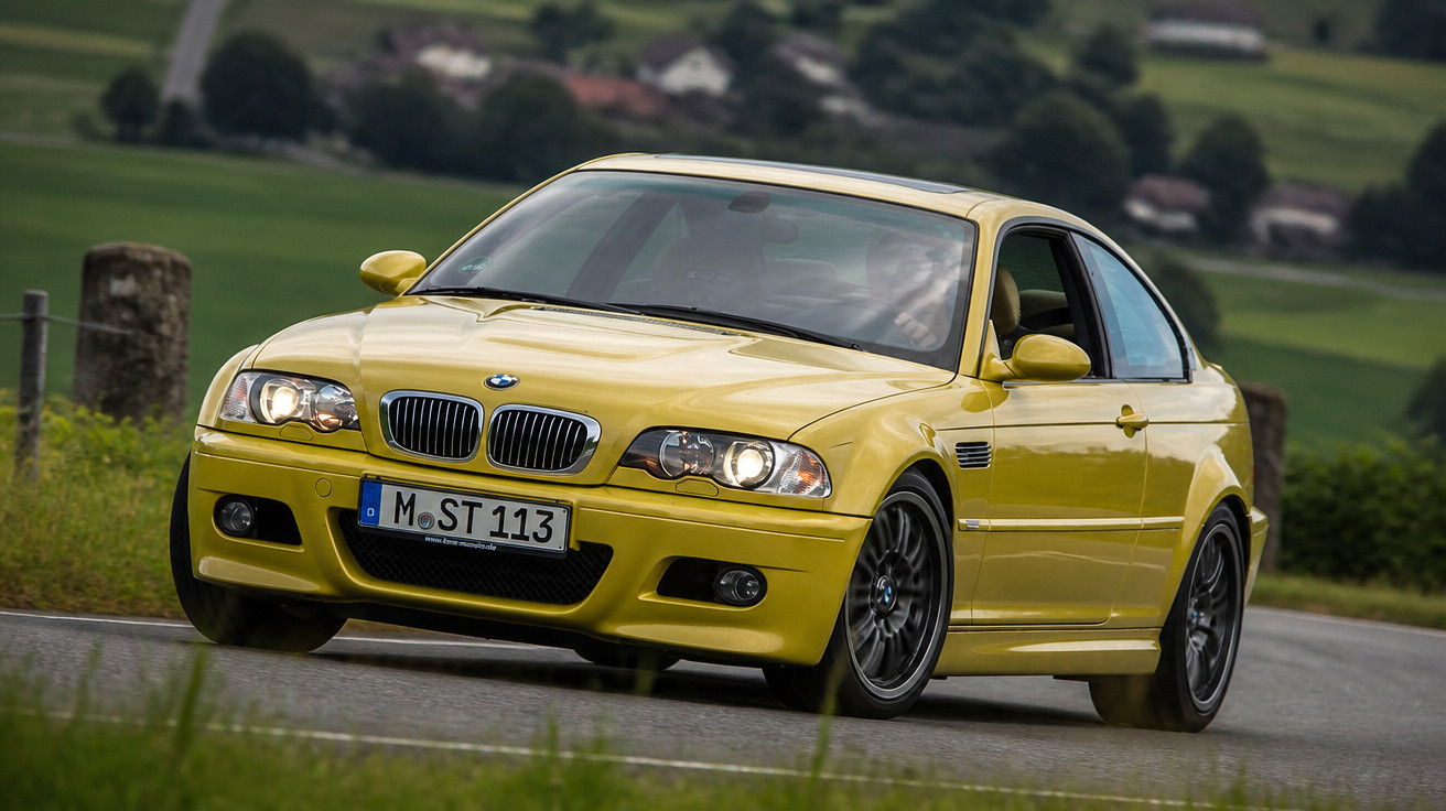 Carro esportivo BMW amarelo em curva de estrada rural cercada por área verde e casas ao fundo.