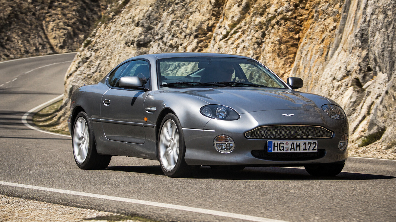 Carro esportivo cinza Aston Martin dirigindo em estrada sinuosa ao lado de paredão rochoso.