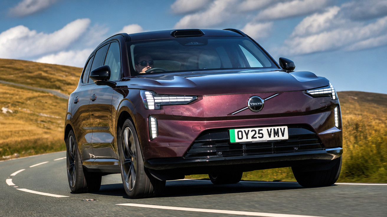 Carro SUV Volvo elétrico vinho em estrada sinuosa com céu azul e nuvens ao fundo.