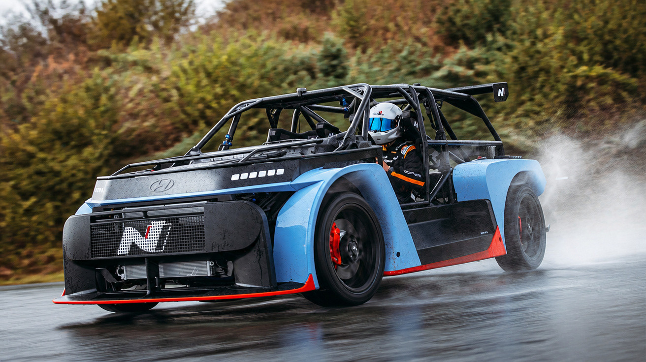 Carro de corrida azul e preto em pista molhada com piloto usando capacete branco.