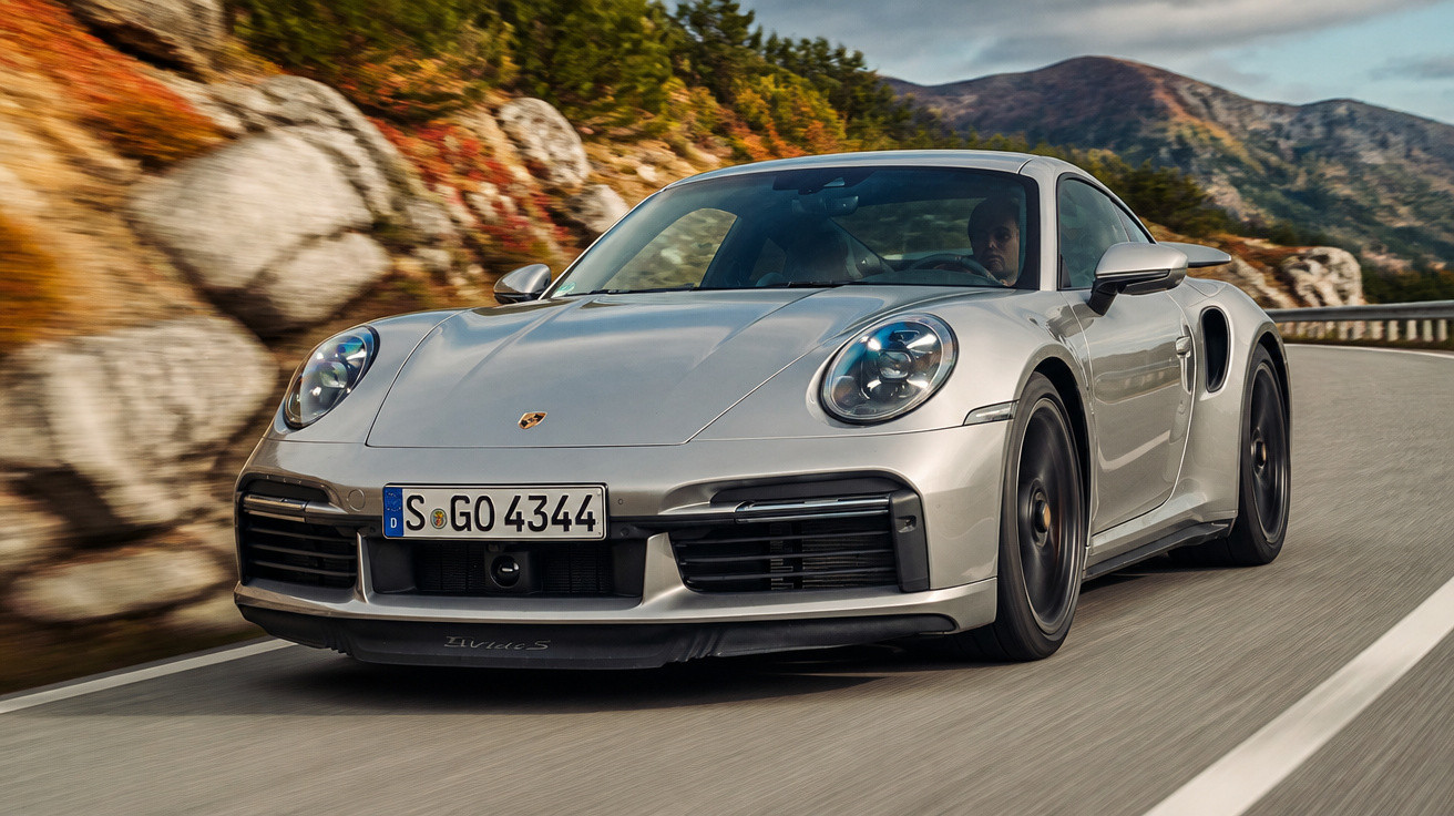 Carro esportivo prata Porsche 911 em estrada montanhosa com paisagem de pedras e árvores ao fundo.
