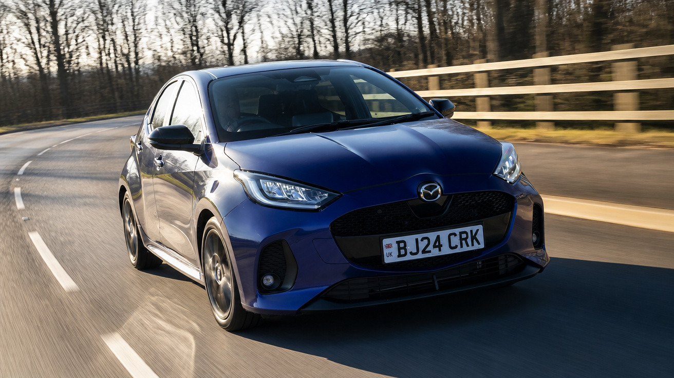 Carro hatchback azul da Mazda em estrada sinuosa cercada por árvores e grade de madeira.